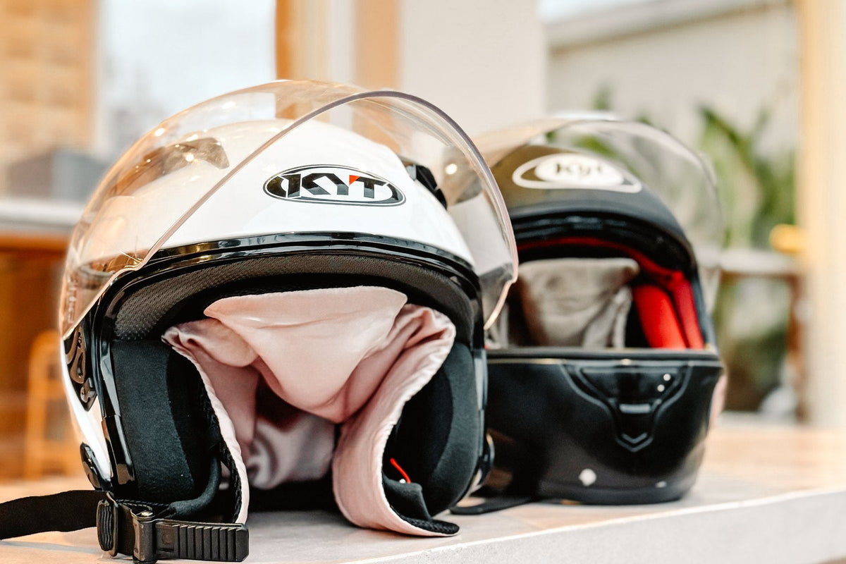 Two motorcycle helmets on a shelf, each fitted with a Silky Days silk insert, one pink and one red.